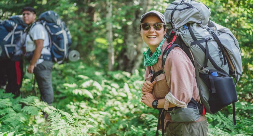 A person wearing a backpack smiles at the camera in a dense green forrest. There are other people wearing backpacks in the background. 